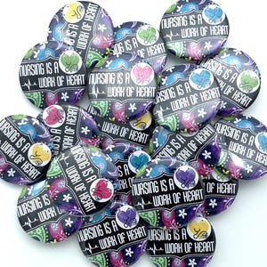 May include: A collection of round buttons with the text "NURSING IS A WORK OF HEART" in white. The buttons have a black background with a paisley design in various colors, including blue, purple, pink, and yellow. A stethoscope graphic is also present.
