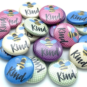 May include: A collection of 12 round, colorful buttons with a bee and the word "Kind" printed on each. The buttons are in various colors, including pink, purple, blue, green, and yellow. The bees are all facing the same direction, and the word "Kind" is printed in black.