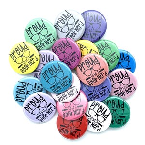 May include: A collection of colorful round buttons with the text "Proud to be a Book Nerd" in black. The buttons are in various colors, including yellow, pink, blue, green, and purple, with a pair of glasses graphic.