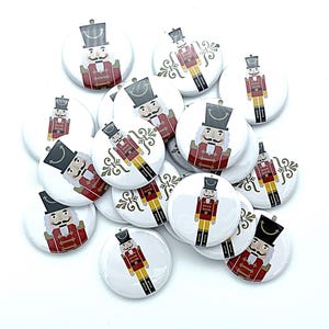 May include: A collection of 15 round, white, button-shaped pins with a red and white nutcracker design. The nutcracker is wearing a red and gold uniform with a black hat. The buttons have a gold trim and a white background.