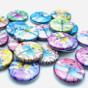 May include: A collection of 15 round buttons featuring colorful dragonfly designs on a white background. The dragonflies are depicted with intricate details and vibrant colors, including blue, green, yellow, and pink.