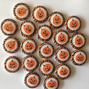 May include: A collection of 18 orange and black Halloween themed buttons with a pumpkin face design. The buttons are round and have a white background with black stripes.
