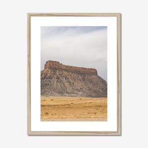 May include: A framed landscape print featuring a mesa in a desert environment. The mesa has a flat top with reddish-brown rock formations. The foreground shows a dry, golden-yellow landscape under a cloudy sky.