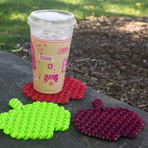 May include: A clear plastic cup with a beverage and a red apple-shaped coaster. Two additional apple-shaped coasters in green and burgundy are also visible. The cup is on a gray stone surface, with a tree and grass in the background.