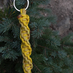 May include: A yellow and black paracord keychain with a silver key ring. The keychain is hanging from a pine tree branch.