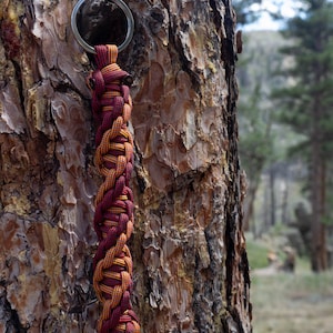 May include: A red and orange paracord keychain with a silver key ring hanging from a tree trunk.