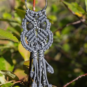 May include: A gray and black macrame owl keychain with black lava rock beads for eyes. The keychain is hanging from a silver key ring.