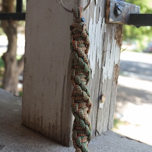 May include: A green and orange paracord keychain with a silver key ring. The keychain is hanging from a wooden post.