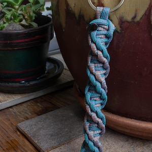 May include: Braided keychain with a silver ring. The keychain is made of interwoven teal, brown, and purple cord. The design is a tight, twisted pattern. The background includes a large brown pot and a small potted plant.