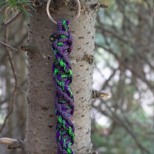 May include: A purple and green paracord keychain with a silver key ring. The keychain is hanging from a tree branch.