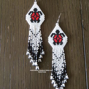 May include: A pair of beaded earrings featuring a white diamond-shaped design with a red and black turtle motif. The earrings have a fringe of white and black beads. The text "#KJ574E" is at the top. The text "©2025 Killikina's Beadwork/Killikina Jung Original Design" is at the bottom.