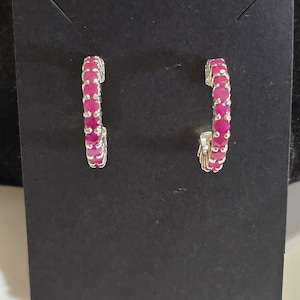 May include: A pair of silver hoop earrings, each lined with vibrant pink gemstones. The earrings are displayed against a dark background, highlighting their color and design. The gemstones are closely set, creating a continuous line of color.
