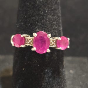 May include: A silver ring featuring three oval-cut, vibrant pink gemstones. The central stone is larger than the two flanking stones. The ring is set against a dark background, highlighting the gemstones' color and sparkle.