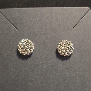 Könnte beinhalten: Ein Paar silberfarbene Ohrstecker. Jeder Ohrring ist ein kleines, kreisförmiges Design, das dicht mit winzigen, facettenreichen Steinen gefüllt ist. Die Ohrringe werden auf einer dunkelgrauen Oberfläche präsentiert.