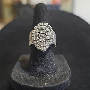 May include: A silver-colored ring with a textured, geometric design. The ring is displayed on a black ring holder. The ring's design features a series of small, irregular shapes, creating a rugged, stone-like appearance. The ring is a statement piece.