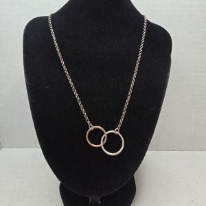 May include: A necklace featuring a silver-toned chain with two interlocking circular pendants. One pendant is a metallic rose gold color, and the other is a polished silver color. The chain consists of small, uniform circular links connected together.