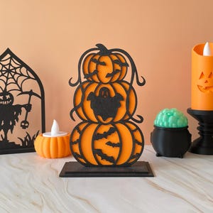 May include: A decorative tabletop ornament featuring three stacked orange pumpkins with black cut-out overlays. The top pumpkin has bat silhouettes, the middle has a ghost shape, and the bottom has more bats. It stands on a rectangular black base.