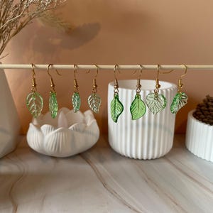 May include: A selection of leaf-shaped earrings with gold-coloured hooks. The earrings are made of translucent green material, each with a unique leaf design. They are displayed on a wooden dowel, with white decorative vases in the background.