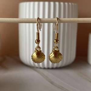 May include: A pair of gold-coloured seashell earrings. The earrings are suspended from gold-coloured hooks and feature a detailed, textured seashell design. The earrings are displayed against a neutral background.