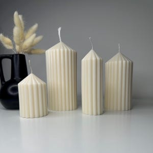 May include: Four ivory-coloured, ribbed pillar candles of varying heights are arranged on a white surface. A black vase with dried flowers is to the left. The candles have a conical top and a wick.
