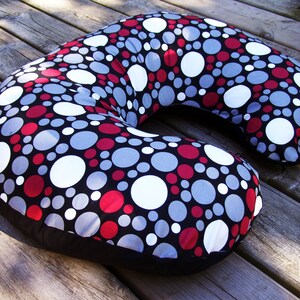 May include: A C-shaped nursing pillow with a black base and a patterned top. The pattern features white, red, and gray circles on a black background. The pillow is designed for comfort and support.