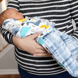 May include: A baby swaddled in a white and blue dinosaur print swaddle blanket. The blanket has a blue and white plaid pattern on the reverse side.