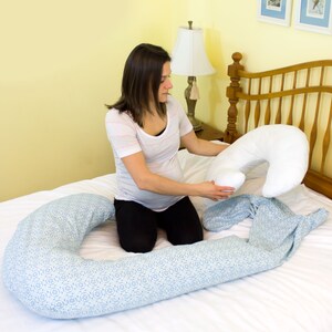 May include: A pregnant person sits on a bed with a white and blue patterned pregnancy pillow. They are holding a white pregnancy pillow in their hands.