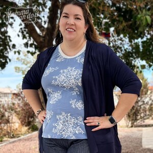 May include: A woman wearing a blue floral print top with a navy blue cardigan. She is standing in front of a green tree.