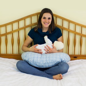 May include: A woman in a teal turtleneck sweater is sitting on a bed with a baby in her arms. The baby is wearing a white and blue onesie and a white hat. The woman is holding the baby on a blue and white patterned nursing pillow.