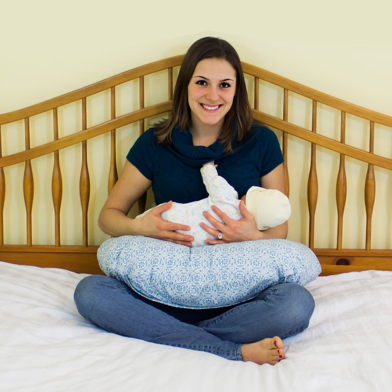 May include: A woman in a teal turtleneck sweater is sitting on a bed with a baby in her arms. The baby is wearing a white and blue onesie and a white hat. The woman is holding the baby on a blue and white patterned nursing pillow.