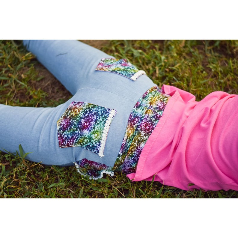 May include: Blue denim pants with rainbow sequin patches on the back pockets. The pants are lying on a bed of green grass.