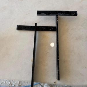 May include: Two black metal T-shaped shelf brackets with a smooth finish. Each bracket features a horizontal top bar with four pre-drilled circular mounting holes and a long vertical support arm extending downward from the center of the top bar.