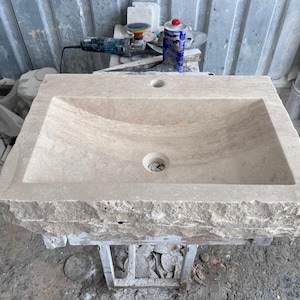 Op de afbeelding: Een rechthoekige, beige stenen wastafel met een ruwe rand. De wastafel heeft een gebogen bekken en twee afvoergaten. Gereedschap en andere stenen stukken zijn zichtbaar op de achtergrond, wat wijst op een werkplaats.