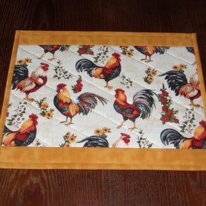 May include: A rectangular quilted placemat with a repeating pattern of colorful roosters, flowers, and foliage on a white background. The placemat has a gold border. The roosters are red, orange, black, and yellow.