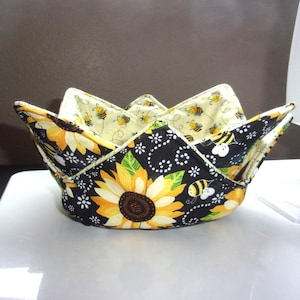 May include: A handmade bowl cozy with a sunflower and bee pattern. The exterior features large yellow sunflowers on a black background with white floral accents. The interior is a light yellow fabric with bee illustrations.