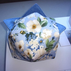 May include: A fabric bowl cozy featuring a light blue background with a floral pattern of white and blue flowers and green leaves. The exterior is quilted with a diamond pattern, and the interior lining is a textured blue fabric.