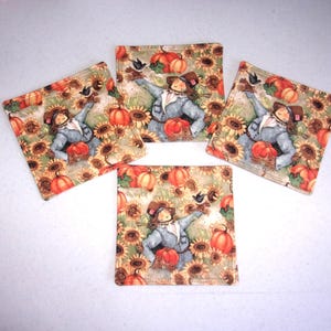 May include: Set of four square fabric coasters featuring a fall harvest theme. Each coaster displays a scarecrow surrounded by sunflowers and pumpkins in shades of orange, yellow, and green. A small black bird is perched on the scarecrow's arm.