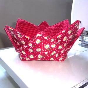 May include: A fabric bowl cozy featuring a vibrant red exterior with a white daisy floral pattern and a contrasting red interior with a small white polka dot print. The bowl cozy has four folded corners and is constructed with visible quilted stitching.