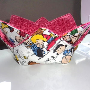 May include: A fabric bowl cozy featuring a white exterior with a colorful Peanuts comic strip print including characters like Snoopy and Charlie Brown. The interior is lined with a bright pink fabric featuring a small, repeating floral pattern.