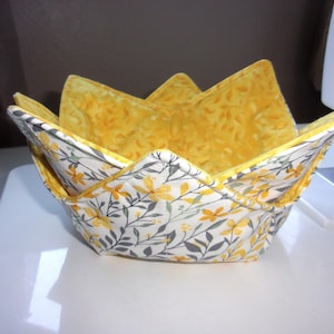 May include: A fabric bowl cozy featuring a white exterior with a yellow and gray floral botanical print and a solid yellow interior. The quilted bowl holder has four pointed corners and is constructed with visible stitching throughout the fabric.