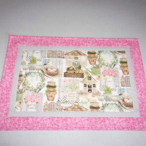 May include: A pink quilted placemat with a floral design featuring a small house, birds, and potted plants. The placemat has a white border and a pink floral trim.