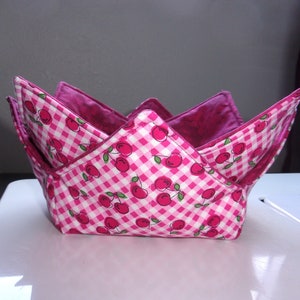 May include: A pink and white gingham fabric bowl with a cherry print. The bowl is folded and has a pink lining.