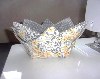 May include: A fabric bowl cozy with a pointed, star-like rim. The exterior features a floral pattern with yellow flowers and gray leaves on a white background, while the interior is lined with gray fabric featuring a white polka dot pattern.