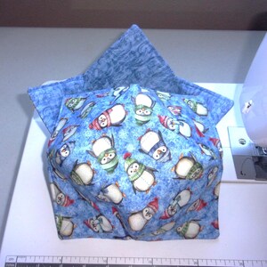 May include: A fabric bowl cozy featuring a repeating pattern of penguins wearing colorful winter hats and scarves against a light blue textured background. The interior lining is a solid blue fabric with a subtle swirling pattern throughout.