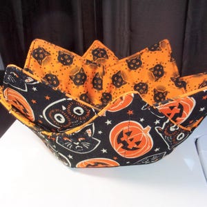 May include: A fabric bowl cozy featuring a reversible design with a black exterior patterned with orange jack-o'-lanterns and black cats, and an orange interior featuring a repeating pattern of small black cat faces and stars throughout the fabric.