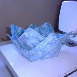 May include: A fabric bowl cozy featuring a light blue and white winter-themed pattern with snowflakes and text that reads Let Snow, Winter Joy, and Wonderland. The square-shaped holder has four pointed corners and is constructed from quilted fabric.