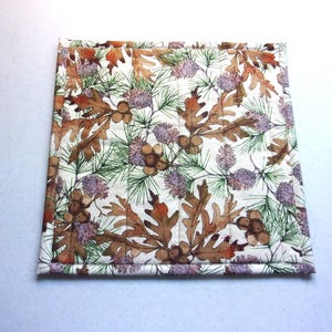 May include: A square quilted fabric potholder featuring a repeating pattern of brown oak leaves, acorns, green pine needles, and purple pinecones on an off-white background. The item has straight stitched lines and a finished fabric border.