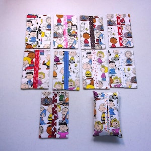 May include: Nine rectangular fabric tissue holders featuring a repeating pattern of Peanuts characters including Charlie Brown, Snoopy, and Lucy. Each holder has a central vertical fabric strip in various colors and patterns like red, blue, and polka dots.