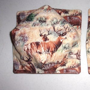 May include: A square fabric bowl cozy featuring a detailed print of brown deer with antlers standing in a field of tall tan grass. The quilted textile accessory has a folded corner design with visible stitching along the edges and a warm color palette.