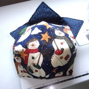May include: A fabric bowl cozy featuring a dark blue background with a snowman pattern, orange noses, red scarves, and yellow stars. The interior is dark blue with white polka dots. A ruler shows the item measures approximately 8 inches wide.